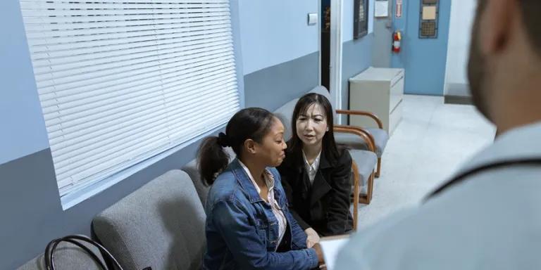 Two women on seats in waiting area with doctor in foreground